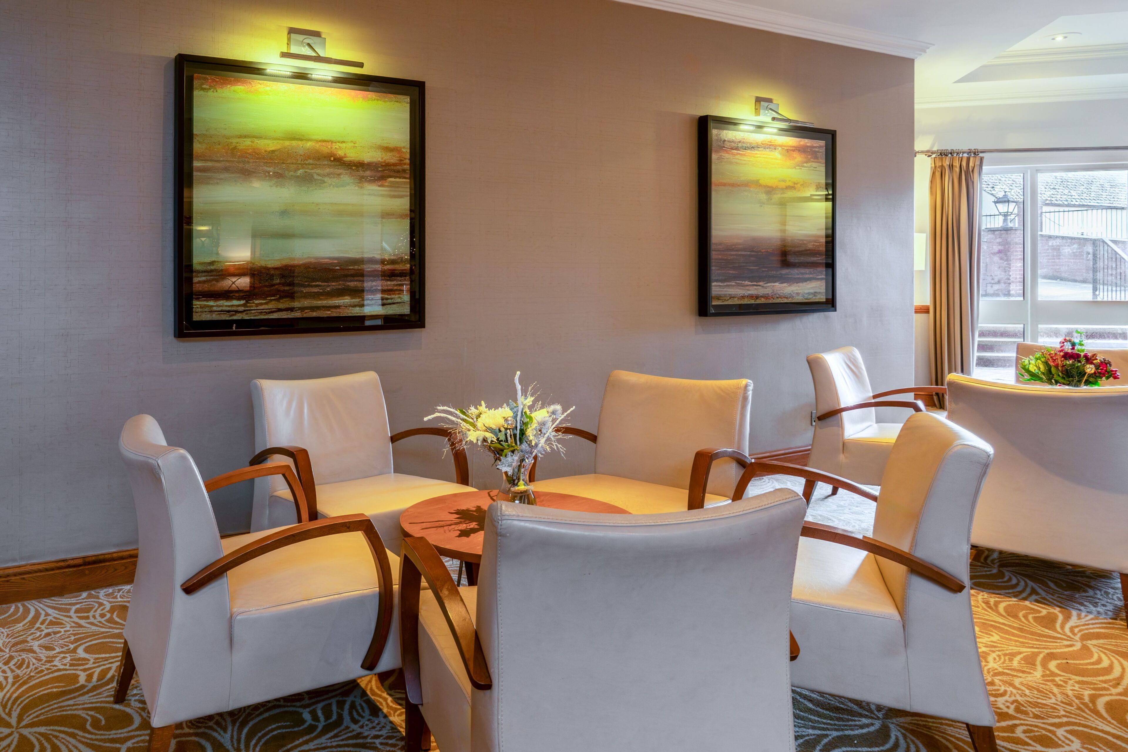 A hotel room with a round table, six chairs, two paintings, a vase with flowers, and a window with curtains.