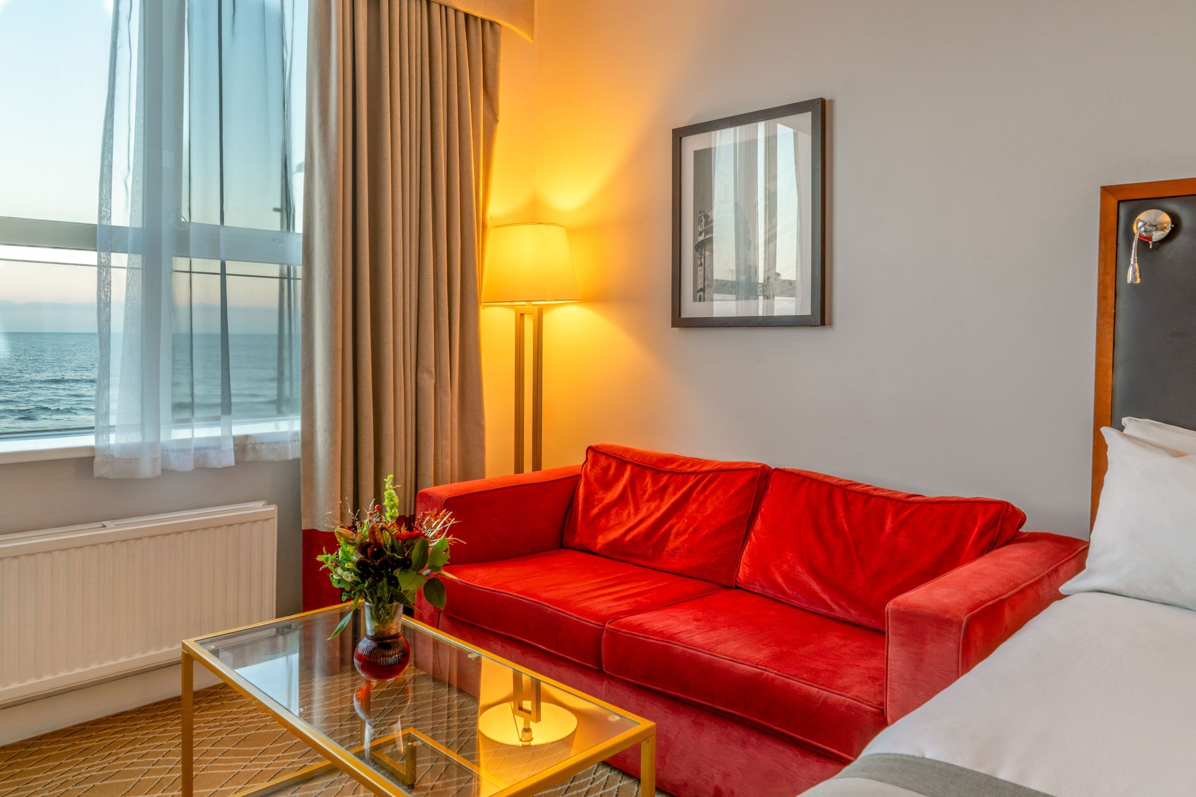 Habitación de hotel con un sofá rojo, una mesa de centro de vidrio, una lámpara, un cuadro en la pared y vista al mar por la ventana.