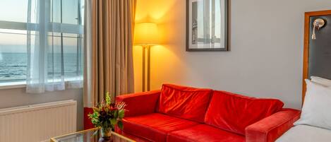 Una camera d'albergo con un divano rosso, un tavolino in vetro, una lampada, un quadro appeso al muro e vista sul mare dalla finestra.