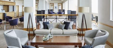 A spacious lobby with a central seating area featuring a sofa and armchairs, a coffee table, and a bar area in the background.