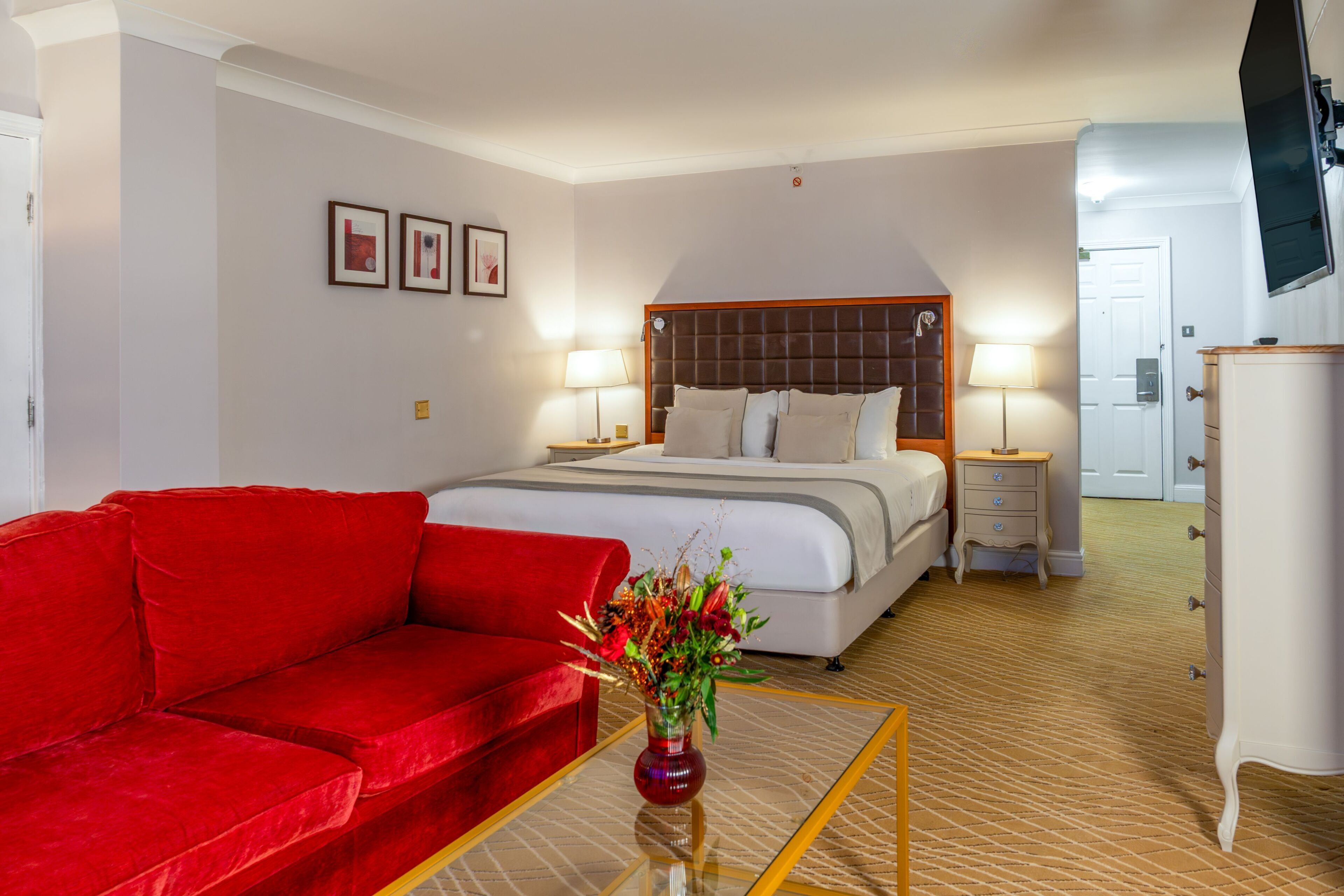 A hotel room with a bed, a red sofa, a TV, a dresser, and framed pictures on the wall.
