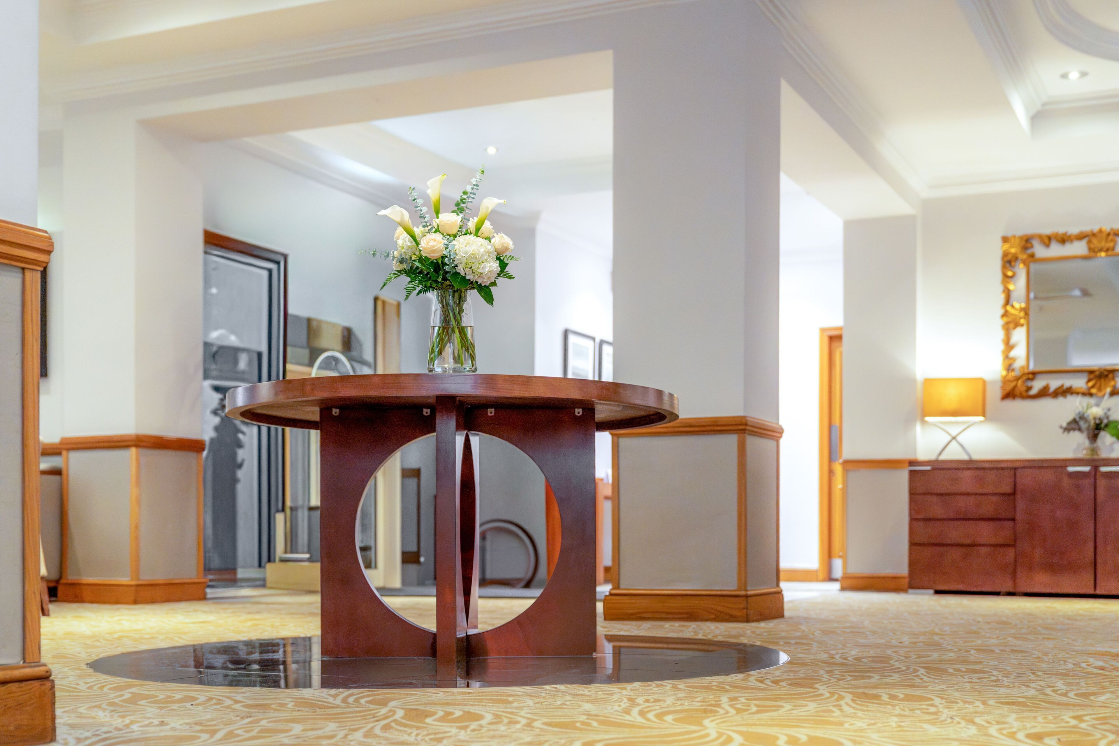 A hotel lobby with a round wooden table, a mirror, and a vase of flowers.