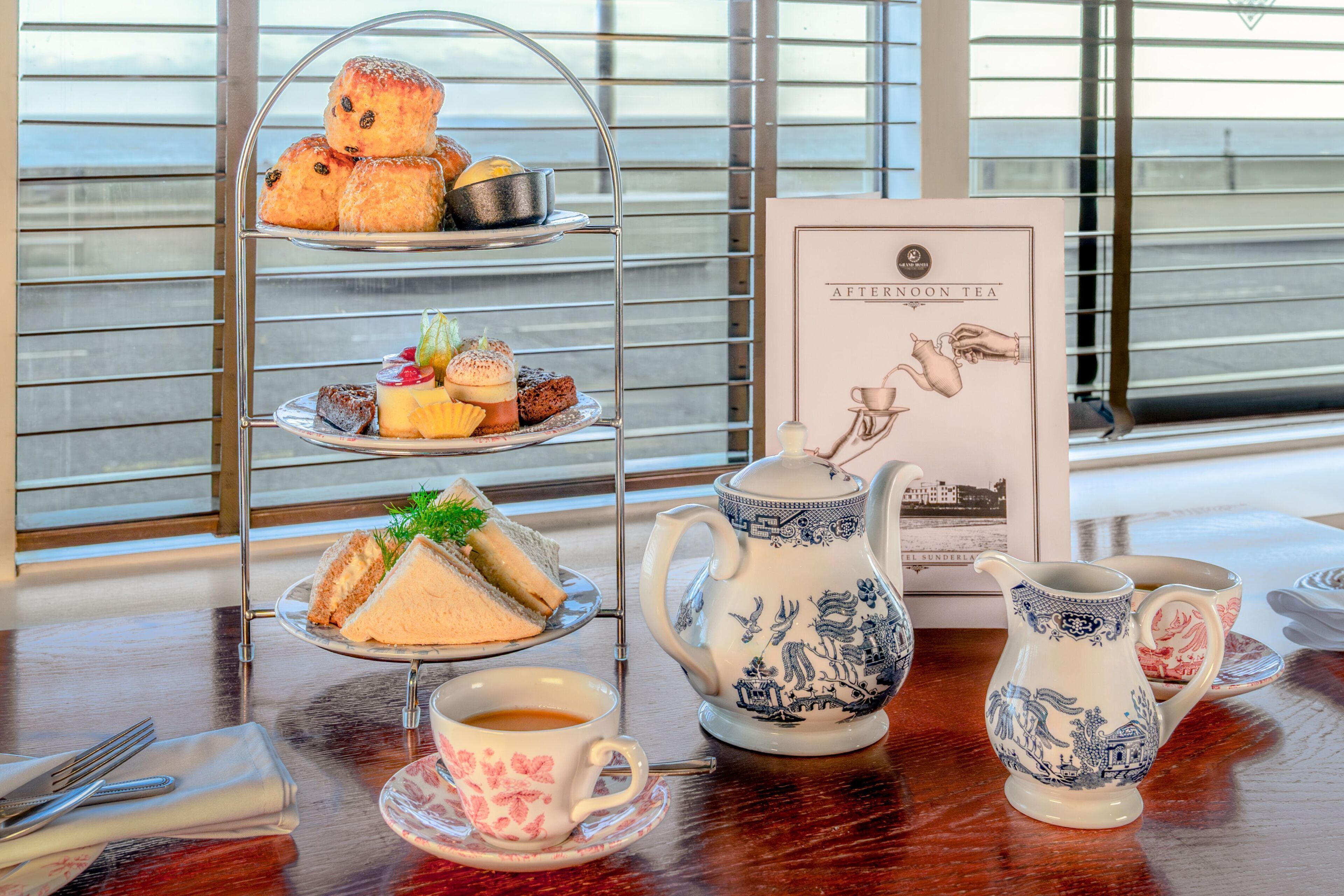 A tiered tray with pastries, a teapot, cups, and a menu on a table by a window.