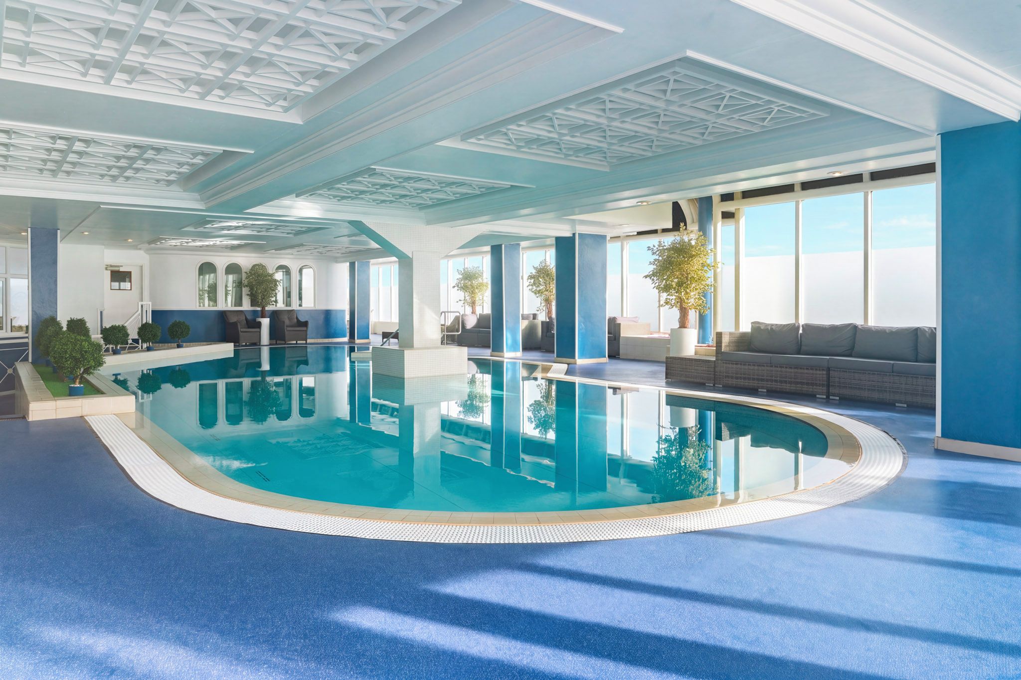 An indoor swimming pool with clear blue water, surrounded by seating areas and potted plants.
