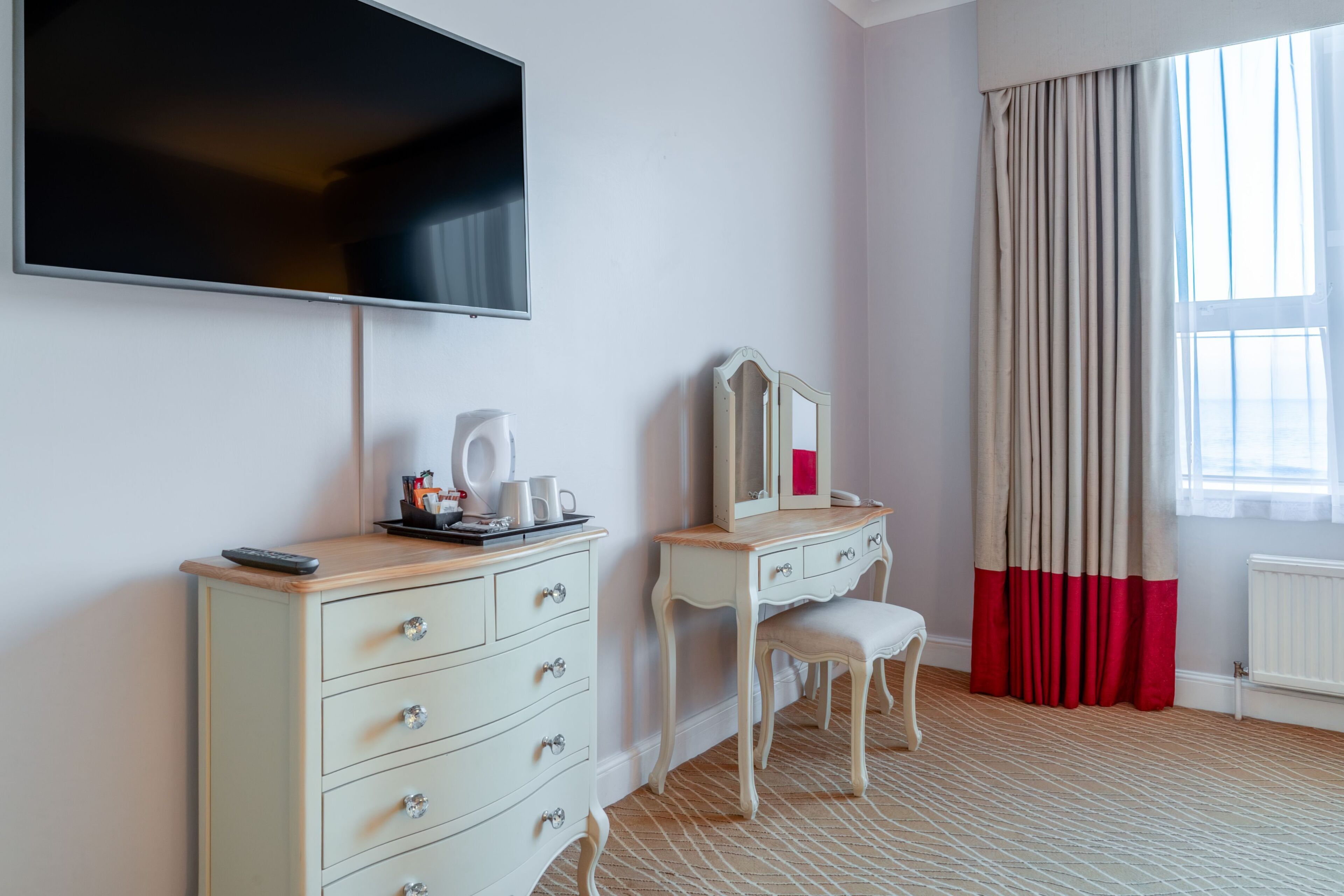 A hotel room with a TV mounted on the wall, a dresser with drawers, a vanity with a mirror, a chair, and a window with curtains.