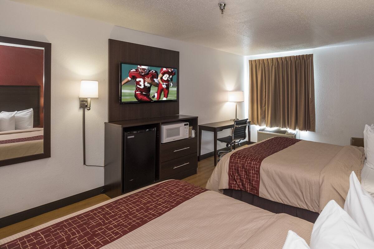 A hotel room with two beds, a TV, a desk with a computer, a microwave, and a window with curtains.