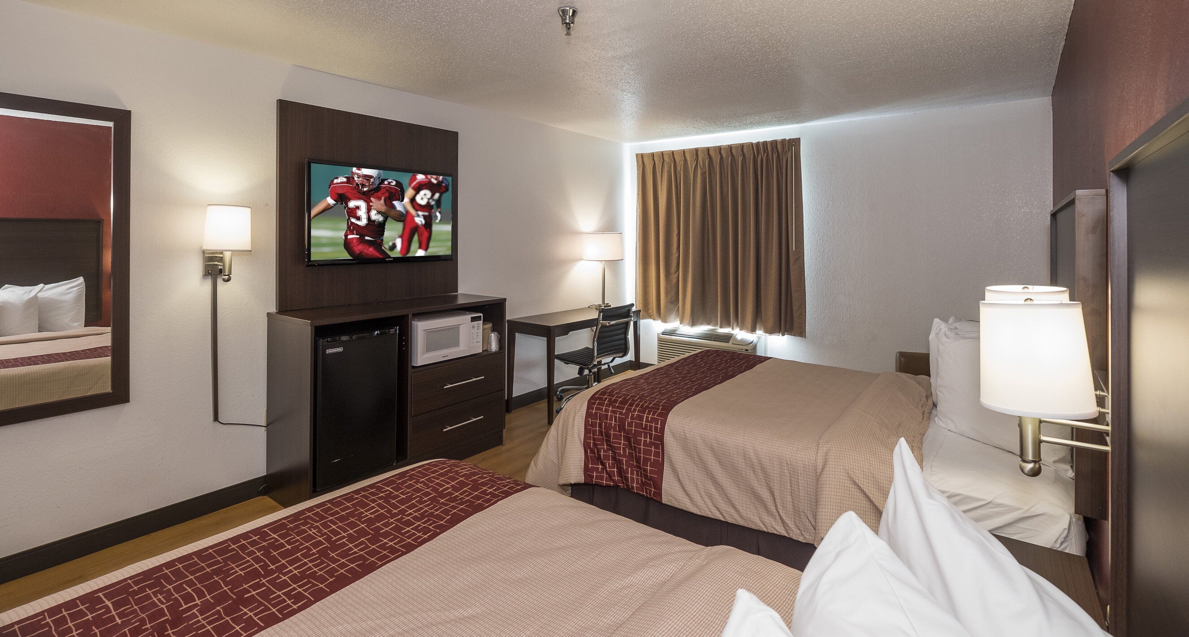 A hotel room with two beds, a television, a desk with a chair, a microwave, and a window with curtains.