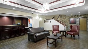 Um hall de hotel com balcão de recepção, um sofá de couro, duas poltronas, uma mesa pequena e uma escadaria que leva a outro andar.