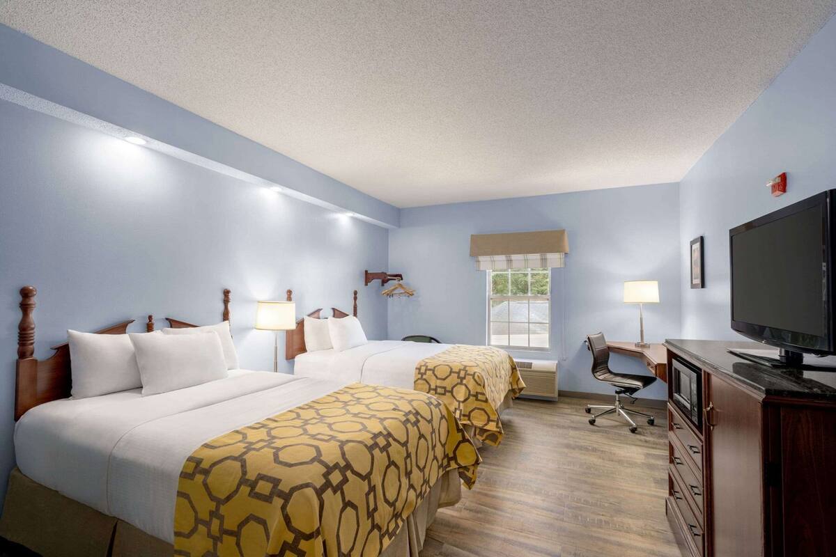Quarto de hotel com duas camas, uma escrivaninha, uma cadeira, televisão e janela.