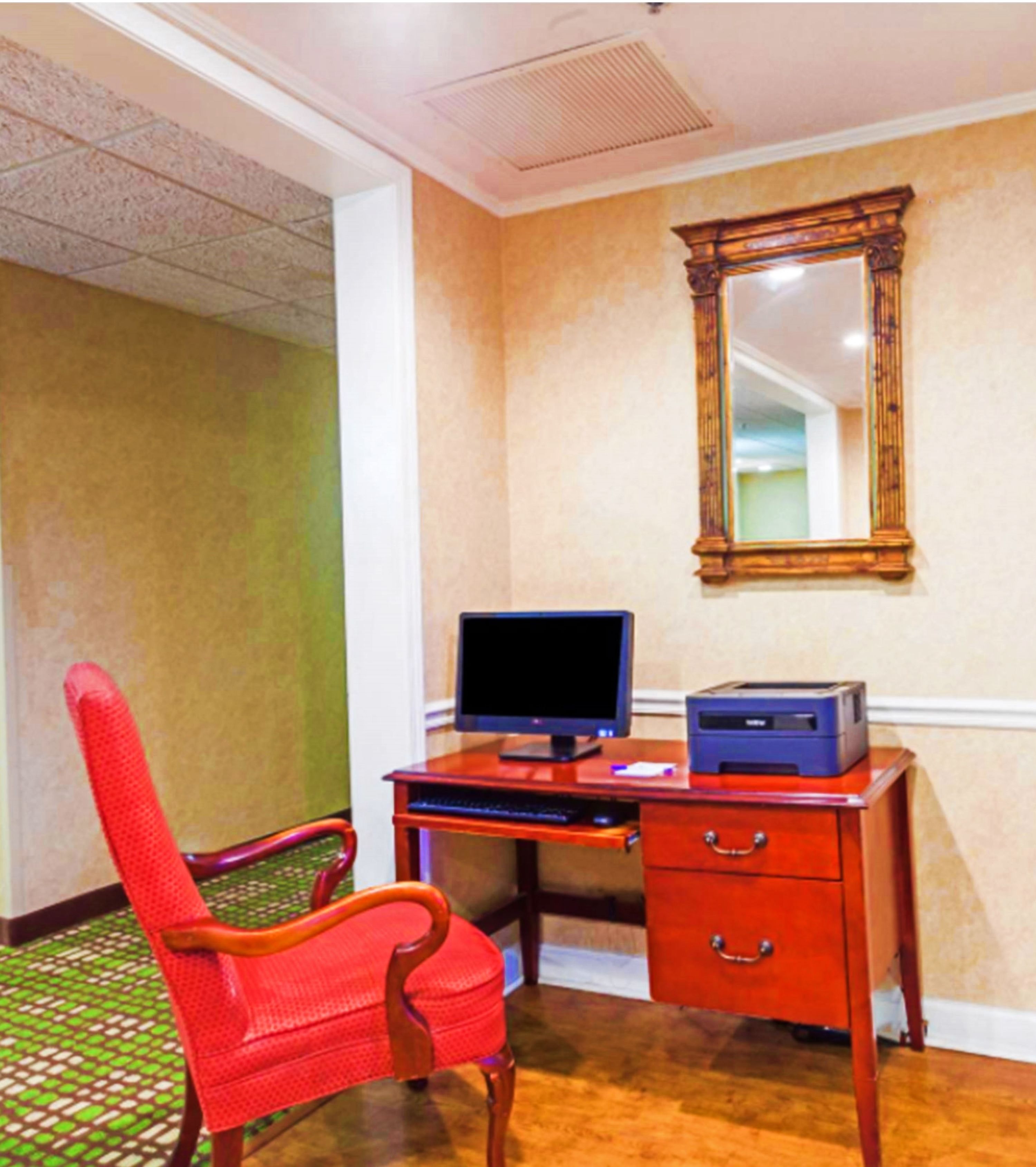 Una habitación con un escritorio de madera, una computadora e impresora. Hay una silla roja y un espejo en la pared.