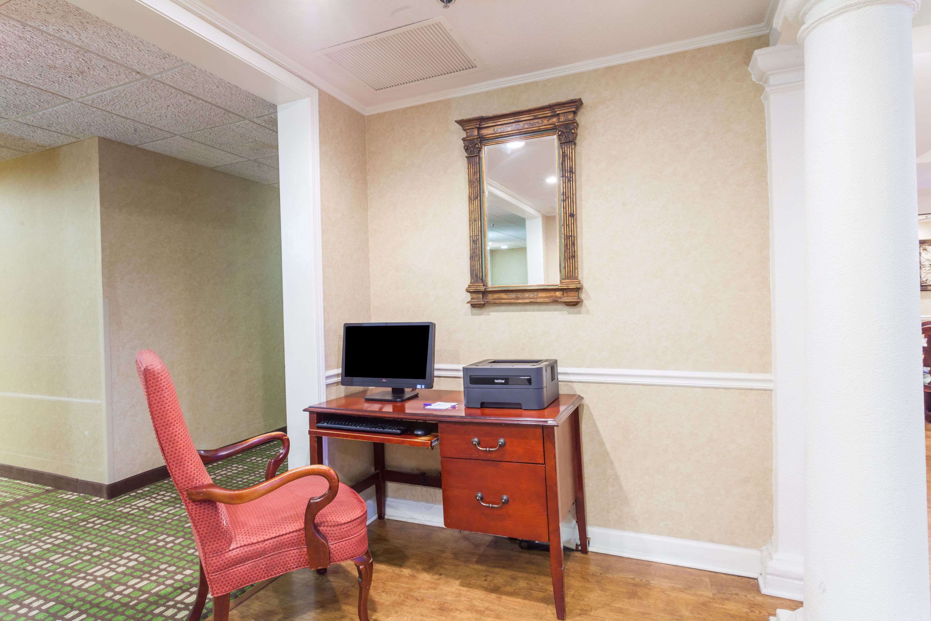 Une chambre d’hôtel avec un bureau en bois, un écran d’ordinateur et une imprimante. Il y a une chaise rouge et un miroir au mur.