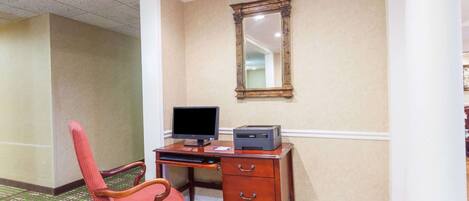 Une chambre d’hôtel avec un bureau en bois, un écran d’ordinateur et une imprimante. Il y a une chaise rouge et un miroir au mur.