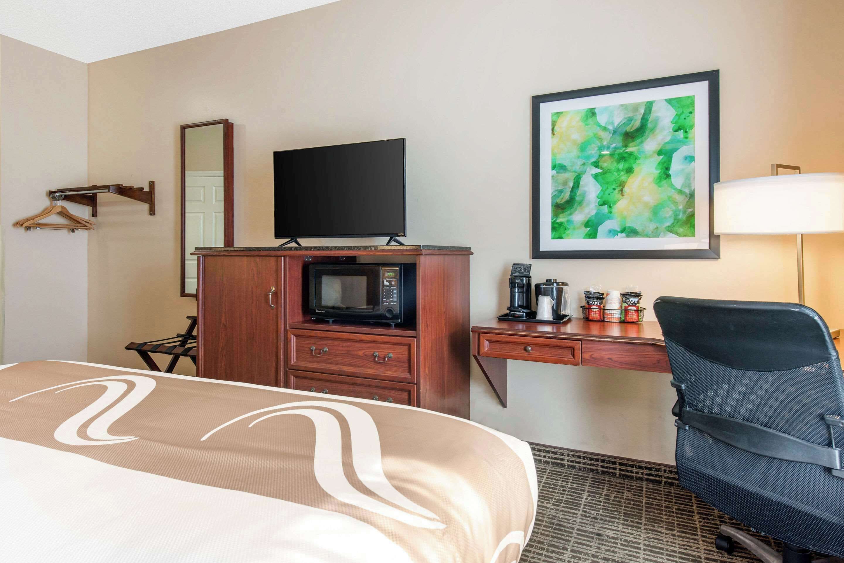 Habitación de hotel con una cama, un escritorio con computadora, microondas, cafetera y un cuadro abstracto en la pared.