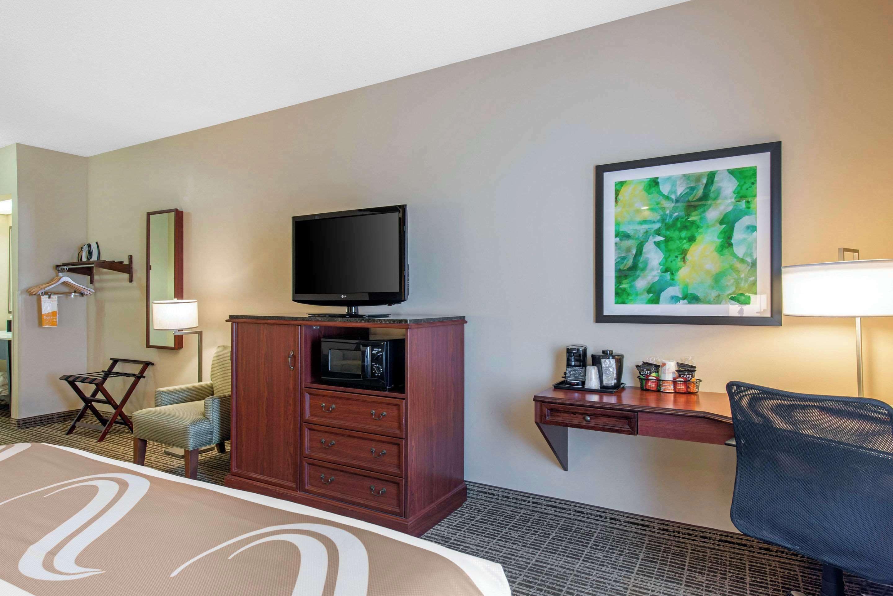 A hotel room with a bed, a TV on a wooden cabinet, a desk with a lamp, a chair, and a painting on the wall.