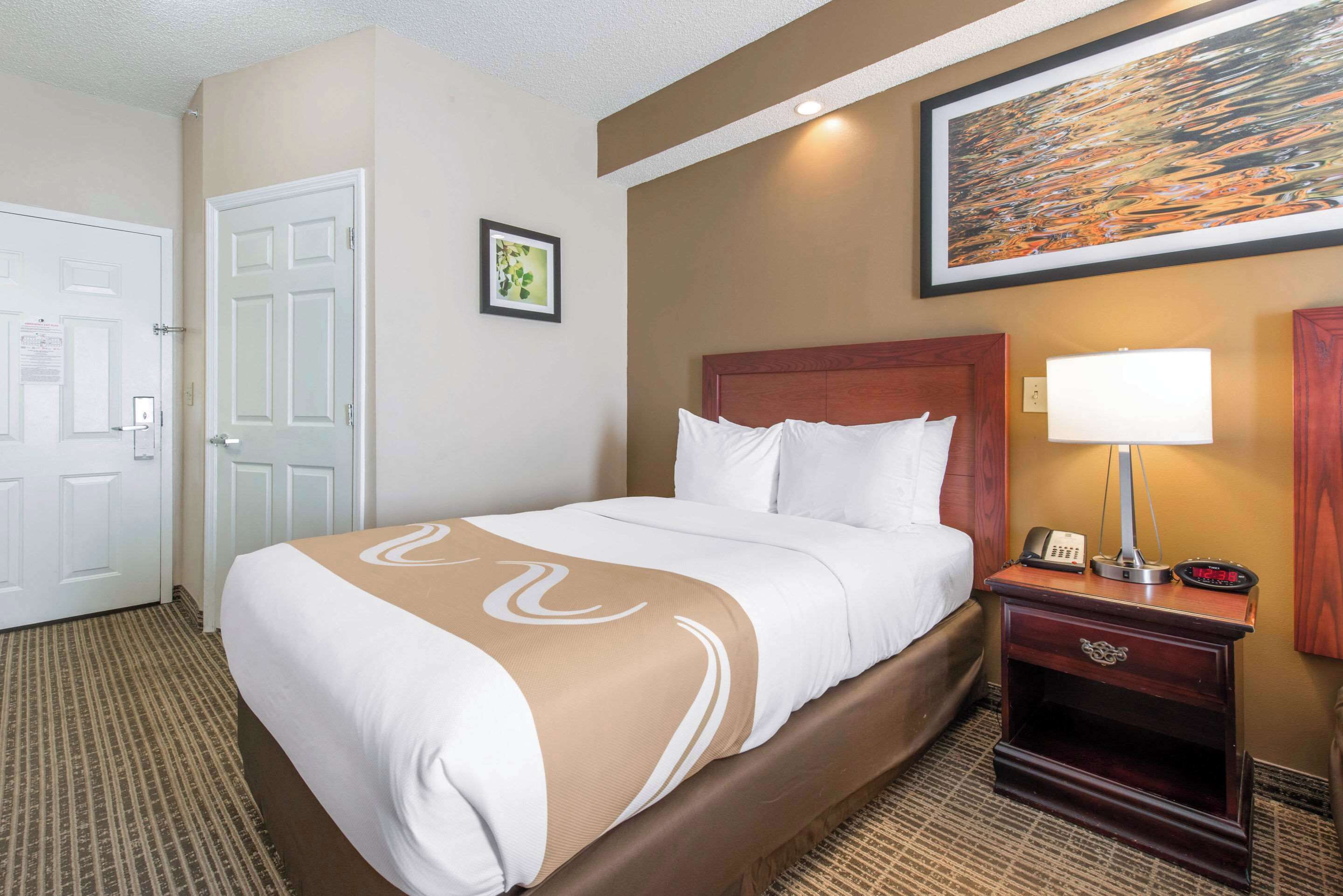 A hotel room with a bed, a nightstand with a lamp, a painting on the wall, and a door.