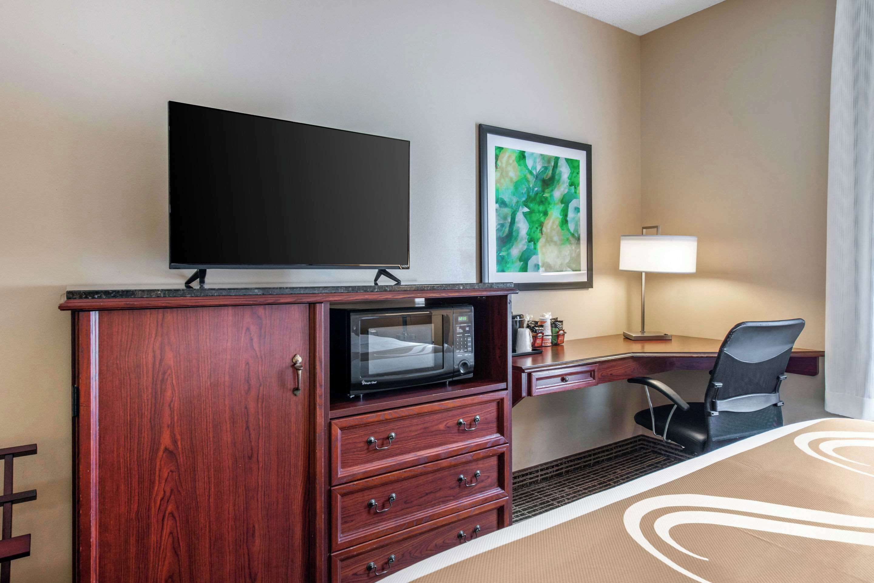 Une chambre d’hôtel équipée d’un téléviseur à écran plat, d’un four à micro-ondes, d’un bureau avec une chaise, d’une lampe et d’un tableau abstrait encadré.