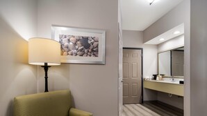 A hotel room with a sofa, a lamp, a framed picture, a door, and a bathroom with a mirror and sink. - Quality Inn Goldsboro (Goldsboro)