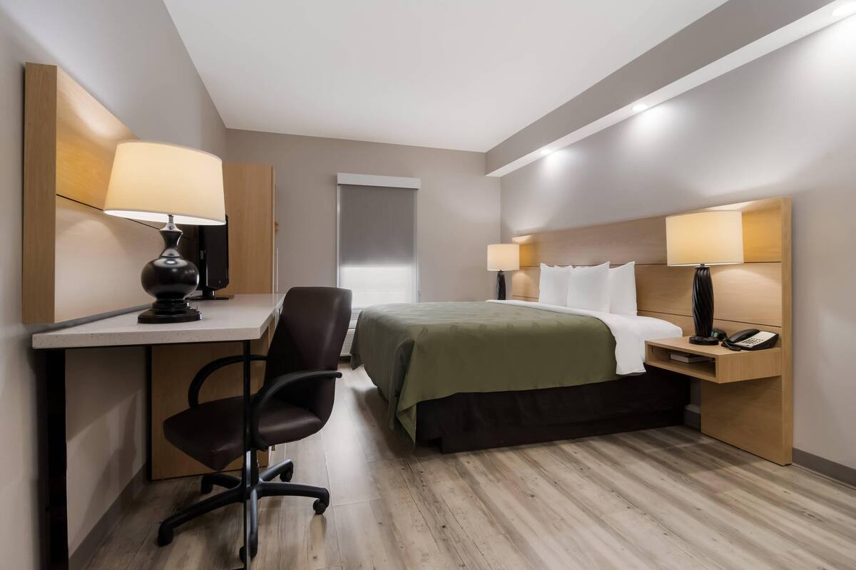 A hotel room with a bed, desk, chair, lamps, and a telephone.
