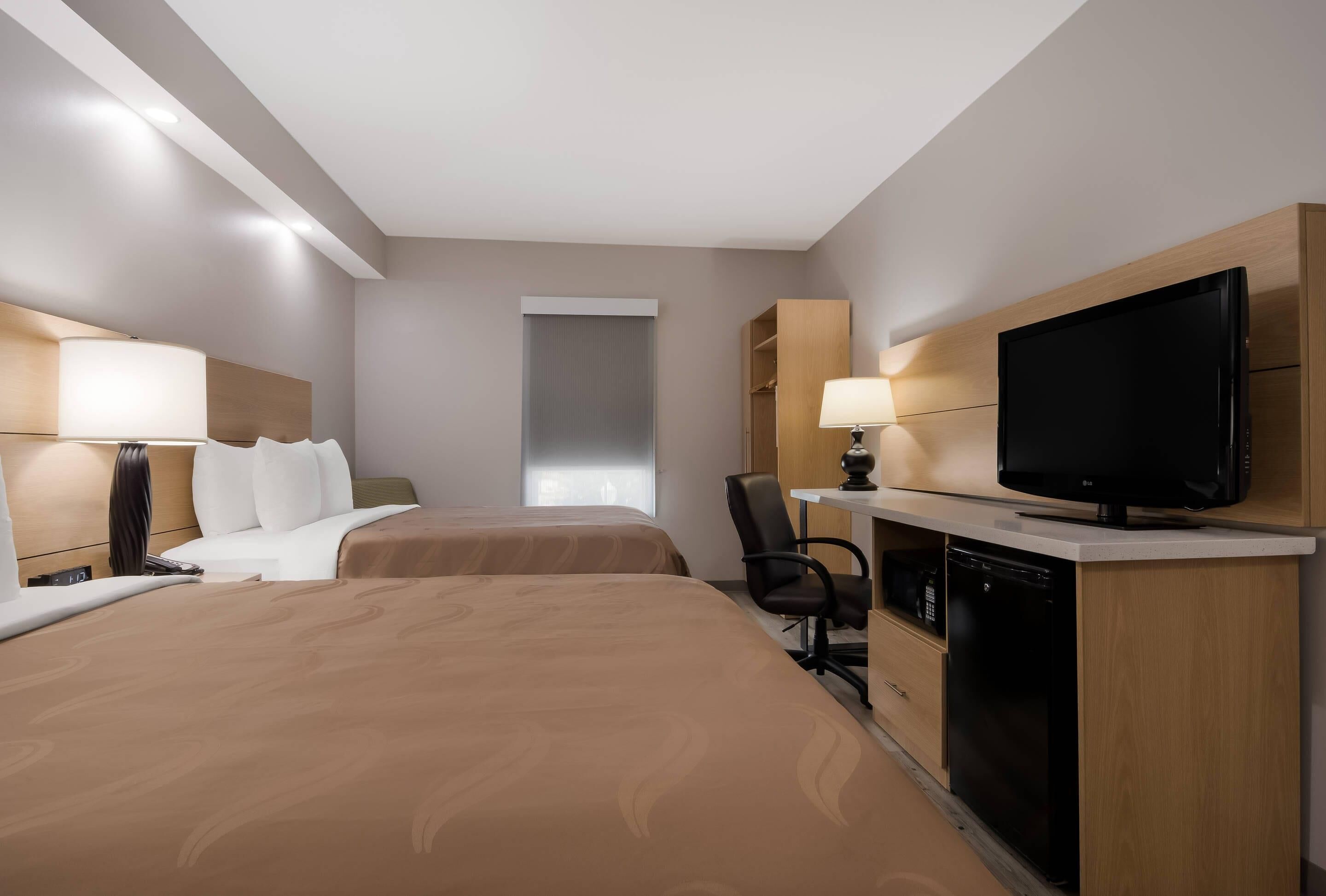 A hotel room with two beds, a desk with a television, a chair, and a small kitchenette.