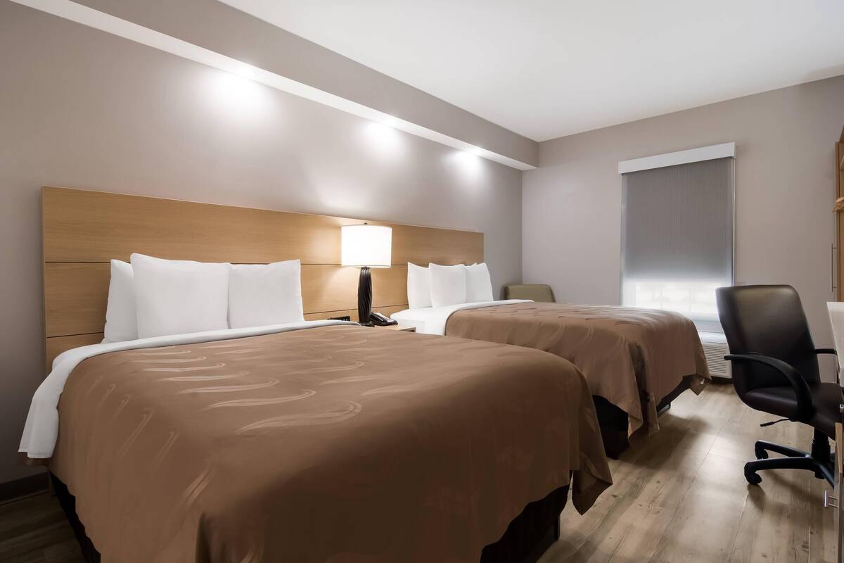 A hotel room with two beds, a desk, a chair, and a window.