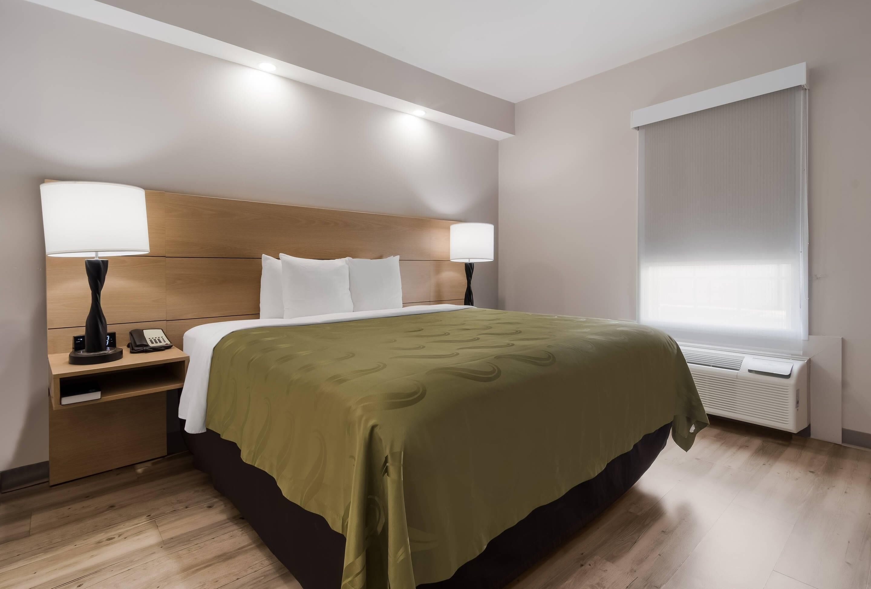 A hotel room with a bed, bedside lamps, a nightstand, a window with blinds, and an air conditioning unit.
