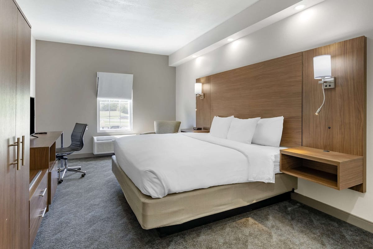 A modern hotel room with a large bed, a desk, a chair, and a built-in shelf.