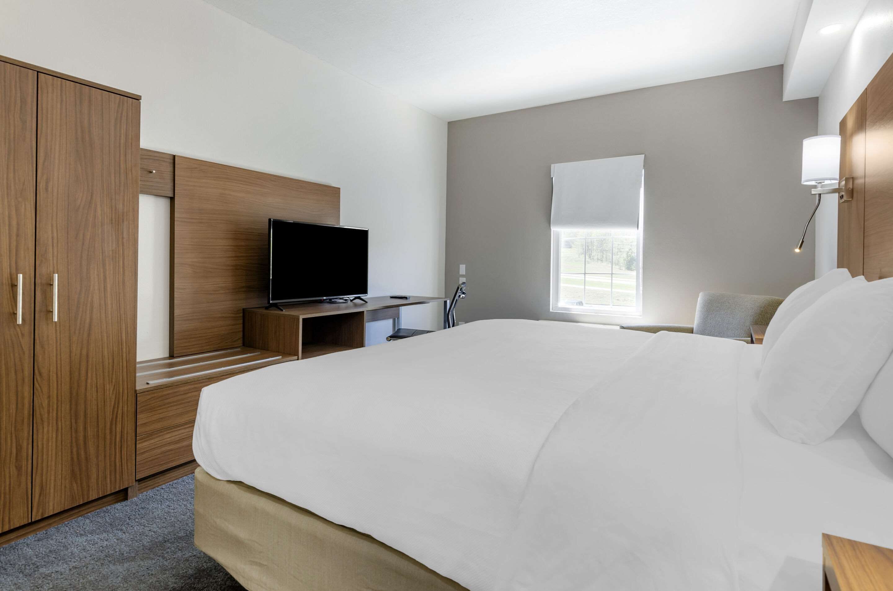 A modern hotel room with a large bed, a TV on a wooden stand, a desk with a chair, and a window with blinds.