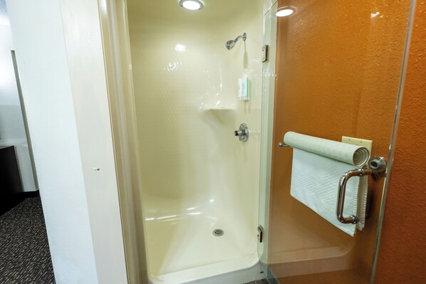 Une cabine de douche fermée par des parois de verre, avec un intérieur blanc, un porte-serviettes avec une serviette et un petit flacon de shampoing.