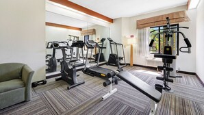 Gimnasio de hotel con equipo de ejercicio, un espejo, una banca y una ventana con cortinas.