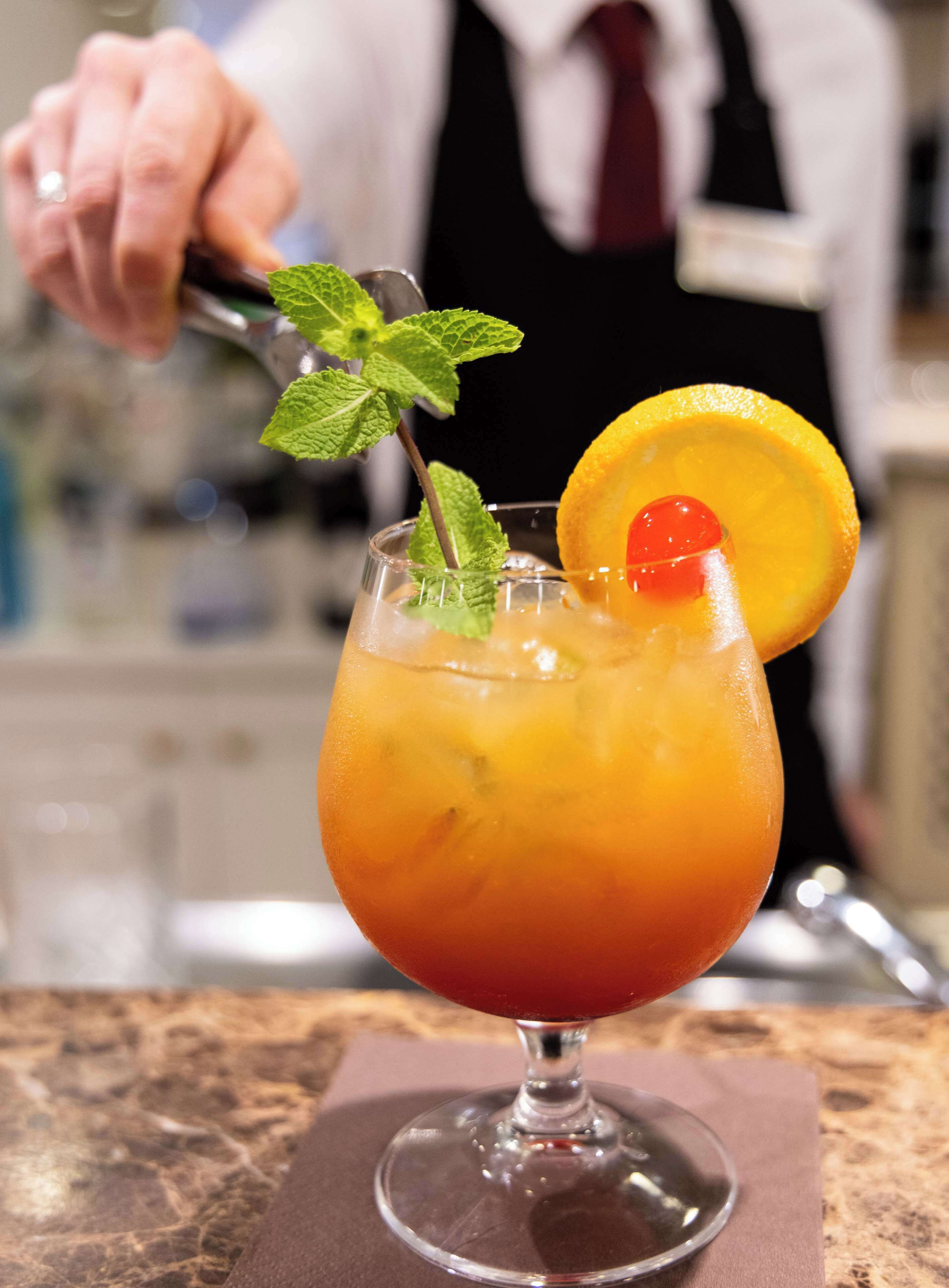 Un cocktail servi dans un verre à pied, agrémenté d’une tranche d’orange et d’une cerise, posé sur le comptoir en marbre du bar.