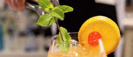 Un cocktail servi dans un verre à pied, agrémenté d’une tranche d’orange et d’une cerise, posé sur le comptoir en marbre du bar.