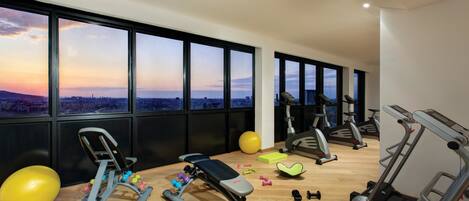 Uma academia com diversos equipamentos de exercício, amplas janelas com vista para a cidade e piso de madeira.