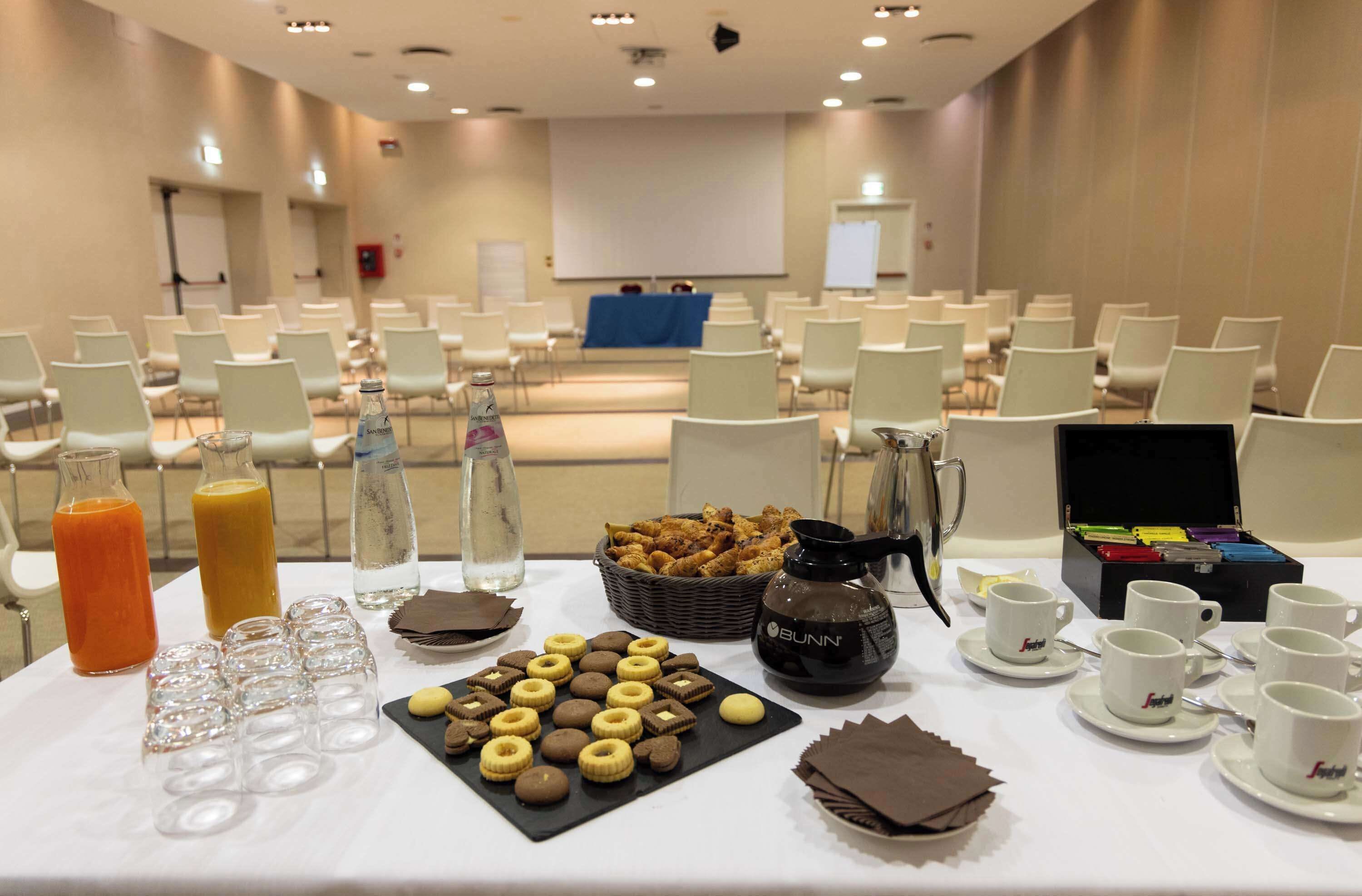 Sala de conferência com fileiras de cadeiras, uma tela de projetor e um buffet com lanches e bebidas.