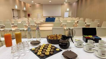 Sala de conferência com fileiras de cadeiras, uma tela de projetor e um buffet com lanches e bebidas.