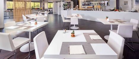 En moderne restaurant med hvide borde og stole, et barområde med et udvalg af vin, og udsigt til det fri.