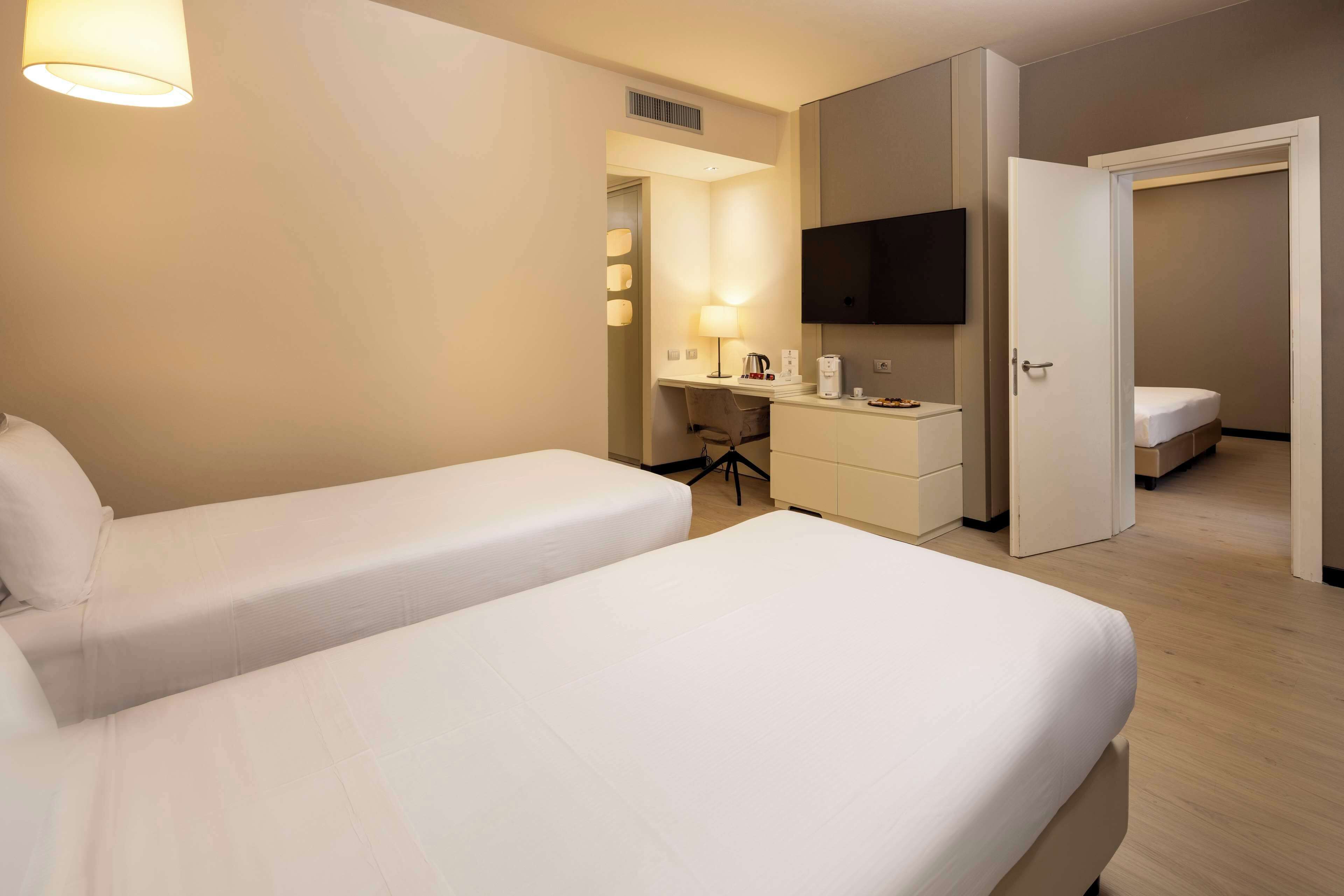 Quarto de hotel com duas camas de solteiro, uma escrivaninha com cadeira, televisão fixada na parede e um guarda-roupa.