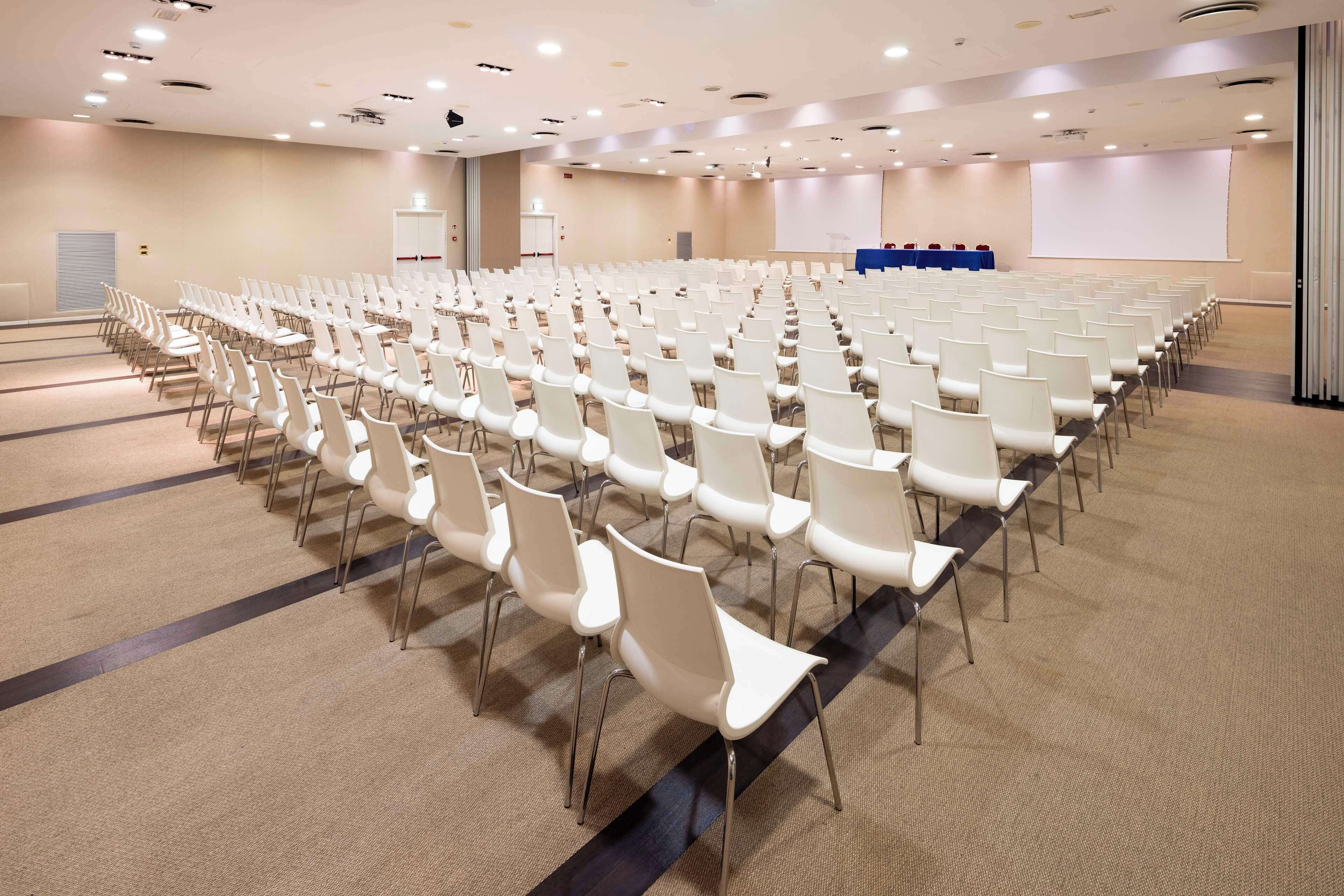 A conference room with rows of white chairs, a carpeted floor, a projector screen, and a presentation area.