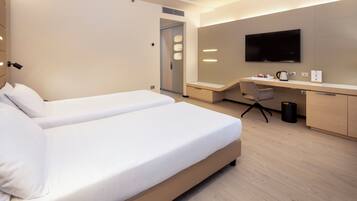 A modern hotel room with two single beds, a desk with a flat-screen TV, and a small seating area.