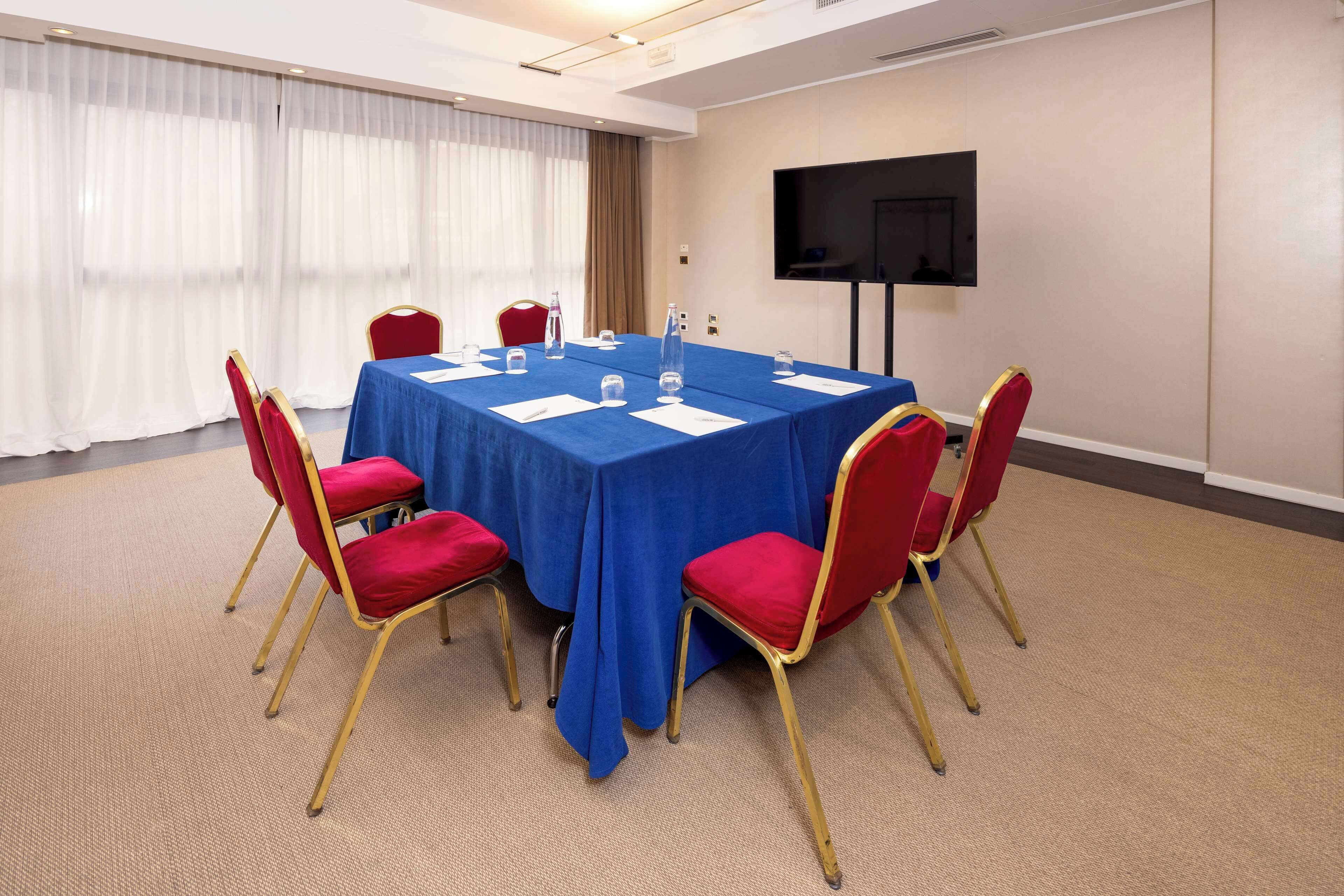 A conference room with a blue table, red chairs, a TV on the wall, and a large window with curtains.