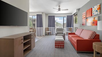Habitación de hotel con televisión de pantalla plana, un sofá, una silla, una mesita, ventilador de techo y una ventana con cortinas.