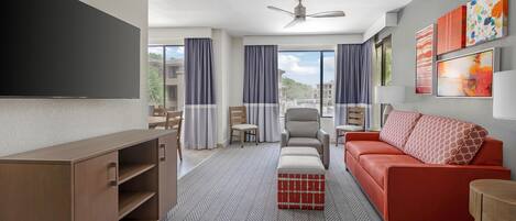 A hotel room with a flat-screen TV, a sofa, a chair, a small table, a ceiling fan, and a window with curtains.