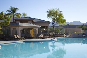 Una piscina con tumbonas y una zona de estar cubierta, rodeada de palmeras y montañas al fondo.