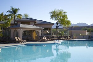 A swimming pool with sun loungers and a covered seating area, surrounded by palm trees and mountains in the background.