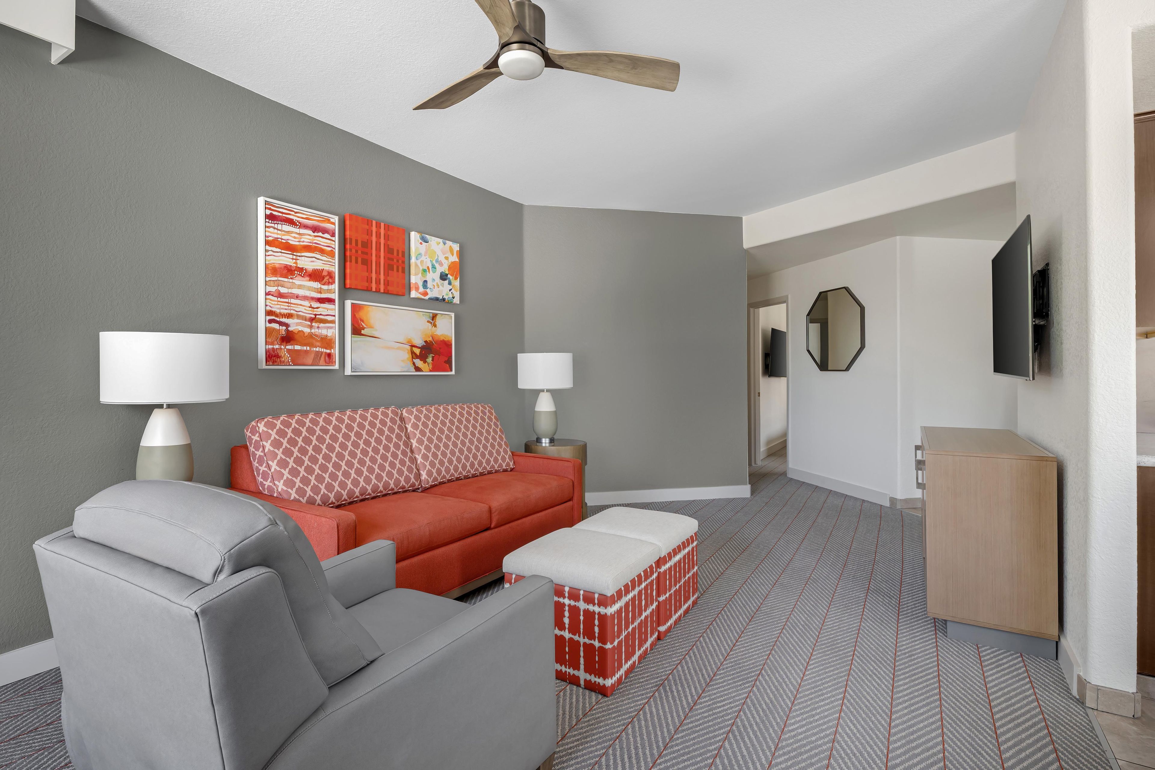 A modern living room with a grey sofa, red ottoman, and a ceiling fan. There are wall-mounted lamps and abstract art.