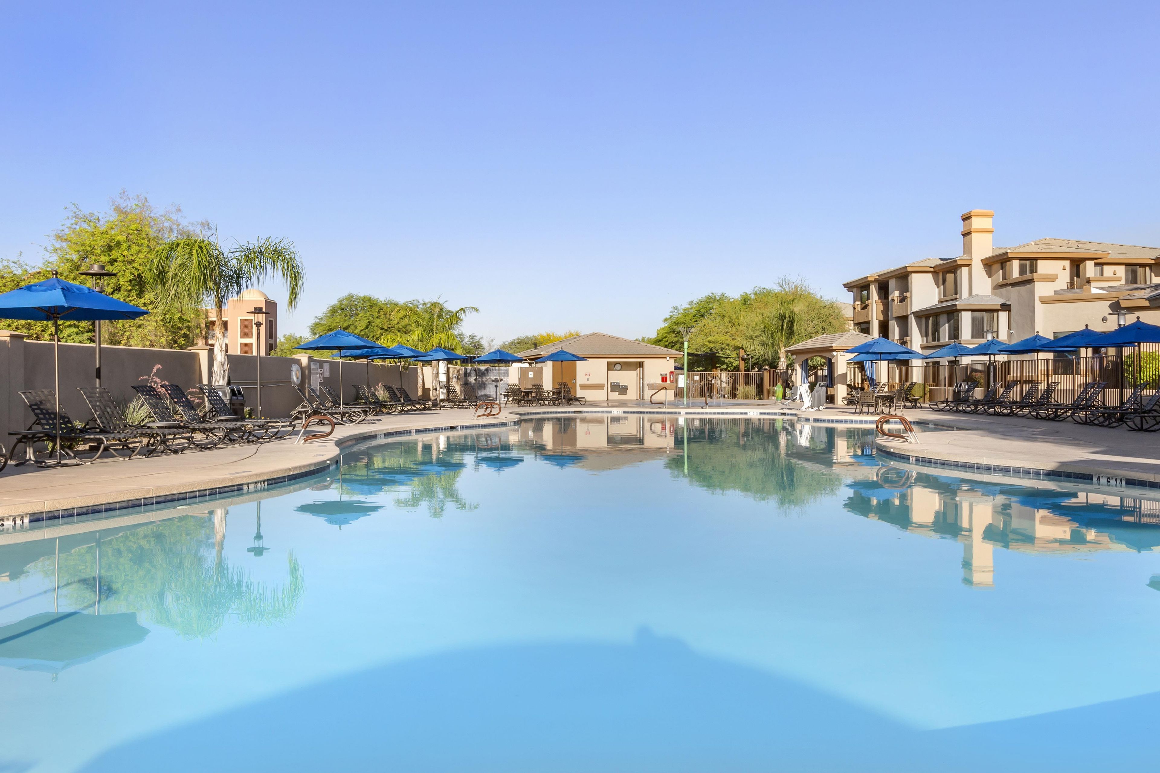 A swimming pool with lounge chairs and umbrellas, surrounded by buildings and trees.