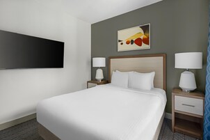 A hotel room with a bed, two bedside tables with lamps, a flat-screen TV mounted on the wall, and a painting above the bed.