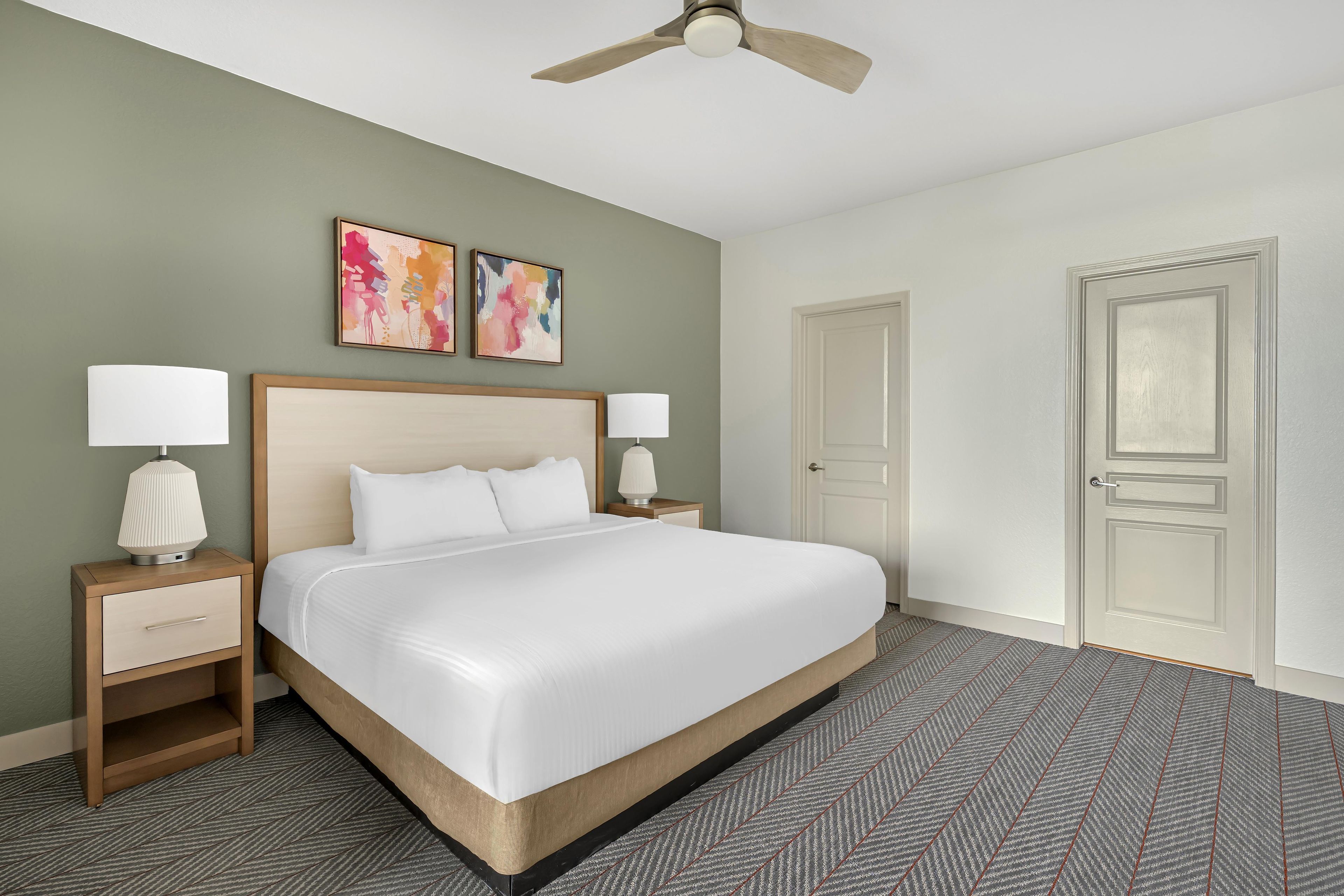 Habitación de hotel con cama, dos mesitas de noche, ventilador de techo y dos cuadros abstractos en la pared.