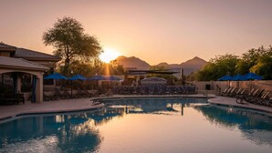 A swimming pool with lounge chairs and umbrellas, surrounded by a building with a covered area and mountains in the background.