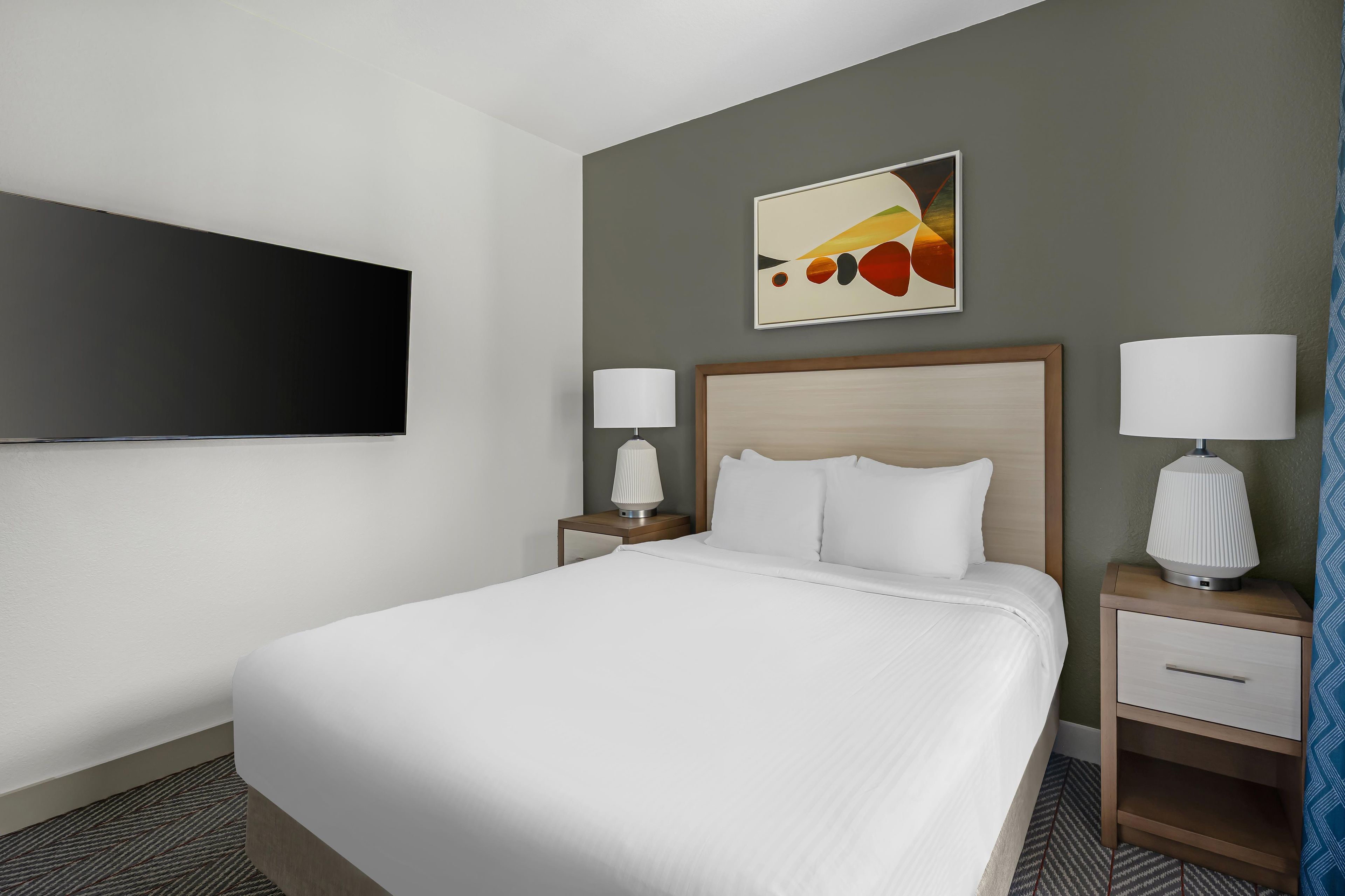 A hotel room with a bed, two bedside tables with lamps, a flat-screen TV mounted on the wall, and a painting above the bed.