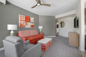 A modern living room with a grey sofa, red ottoman, and a ceiling fan. There are wall-mounted lamps and abstract art.